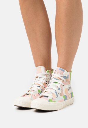 Nogi w wysokich trampkach z płótna w wielokolorowy pastelowy wzór w formie łat oraz białymi sznurowadłami na jednolitym białym tle.