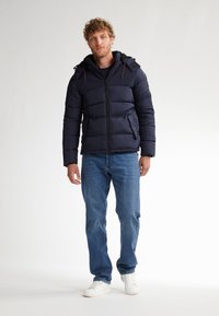 Chaqueta de plumas azul marino con capucha, cierre de cremallera, puños elásticos y bolsillos laterales, combinada con unos vaqueros azules y zapatillas blancas sobre un fondo claro.