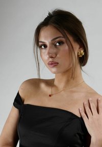 Svart off-shoulder topp, guldfärgad hjärtformad halskedja och en stor guldring med en texturerad design. Slät hud, naturlig sminkning.