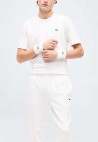 Άνδρας μοντέλο ντυμένο με ολοκίτρινο αθλητικό casual σύνολο με πράσινα λογότυπα κροκόδειλου στη μπλούζα, το παντελόνι και τα βραχιολάκια, στέκεται μπροστά σε απλό φόντο.