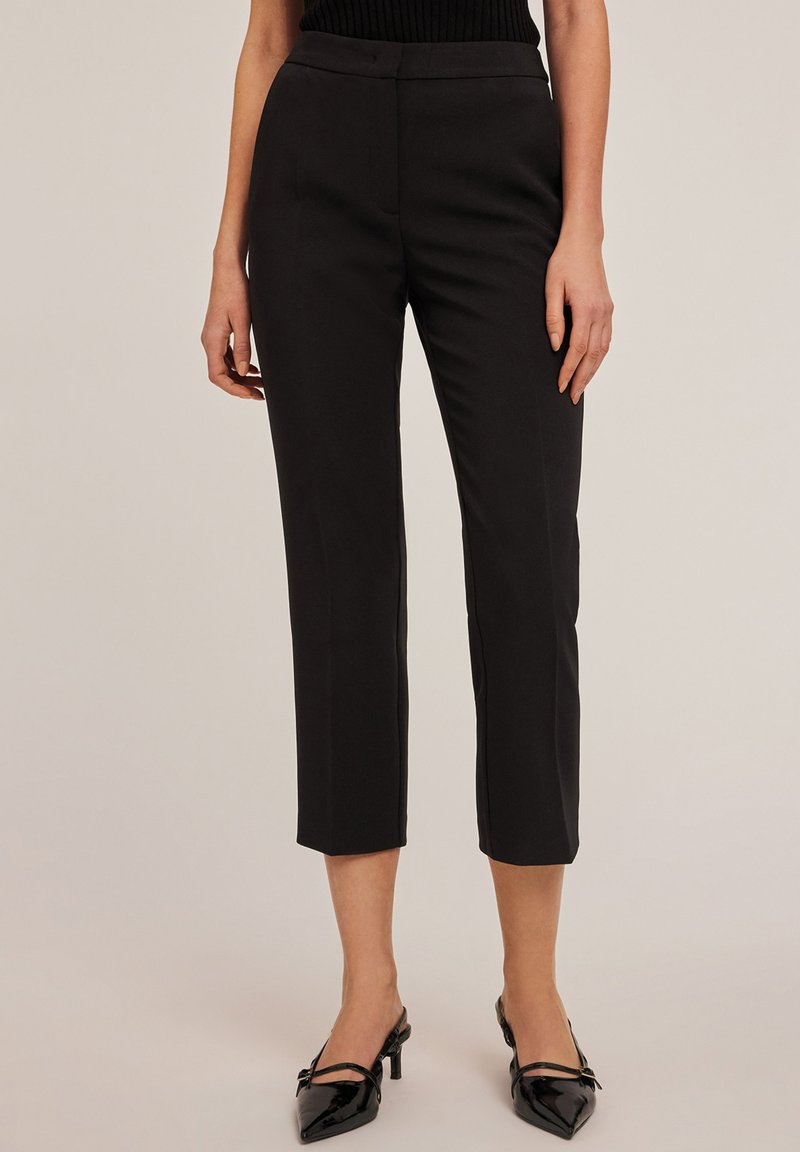 Schwarze, taillierte, cropped Hosen aus glattem Stoff, mit hohem Taillenbund und Seitentaschen, kombiniert mit spitzen schwarzen Absatzmules.