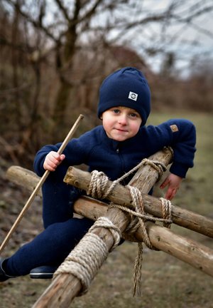 Lapsi tummansininen neule päällään ja pipo päässä istuu rustiikkisella puisella rakenteella, pitelee kädessään oksanpätkää. Rakenteessa on köysikoristeita ja luonnollisia tekstuureja.