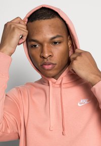 Felpa Nike di colore rosa chiaro, con tasca frontale, cappuccio con coulisse regolabile e logo bianco. Tessuto morbido in misto cotone.