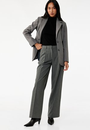 Grijze blazer met twee knopen, zwarte coltrui en grijze wijde broek. Leren enkellaarsjes met puntige neus. Glad textuur, op maat gemaakte pasvorm.