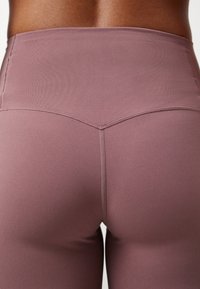 Gros plan sur le bas du dos et les hanches d'une personne portant un legging taille haute mauve avec un détail de couture en forme de V à la ceinture.