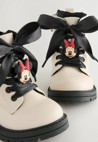 Cremefarbene Knöchelstiefel aus Leder mit breiten schwarzen Satinschnürsenkeln, Gummisohlen und einem Minnie-Maus-Akzent an den Schnürsenkeln.