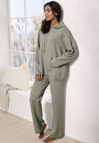 Übergroßer Kapuzenpullover und weite Hose aus sanftem, gedämpftem Grüntuch. Bemerkenswerte Merkmale sind eine Fronttasche und ein Logo-Detail auf der Hose.