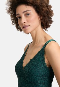 Kant bralette in diep groen met geschulpte randen en dunne bandjes. Kenmerkt zich door een intricaat bloemenpatroon en een gestructureerde stofafwerking.