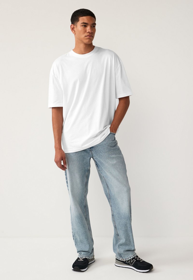 Jeune homme debout, une main dans la poche, portant un t-shirt blanc oversize, un jean bleu clair et des baskets noires sur fond uni.