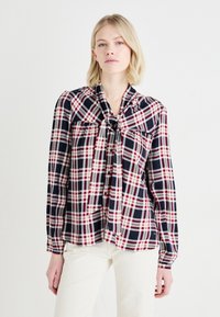 Camicia a scacchi in blu navy, rosso e bianco con dettagli a fiocco al colletto. Maniche lunghe e vestibilità comoda, con chiusura a bottoni davanti.