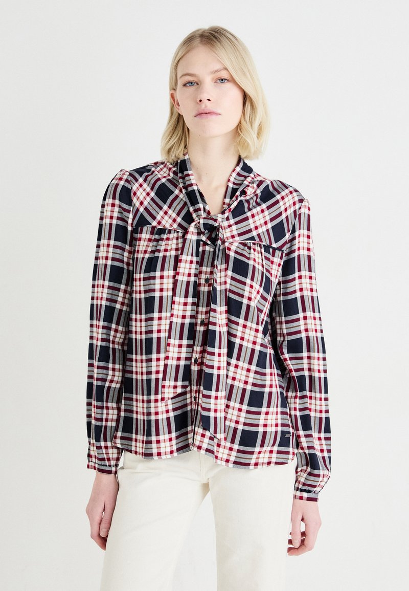 Camicia a scacchi in blu navy, rosso e bianco con dettagli a fiocco al colletto. Maniche lunghe e vestibilità comoda, con chiusura a bottoni davanti.