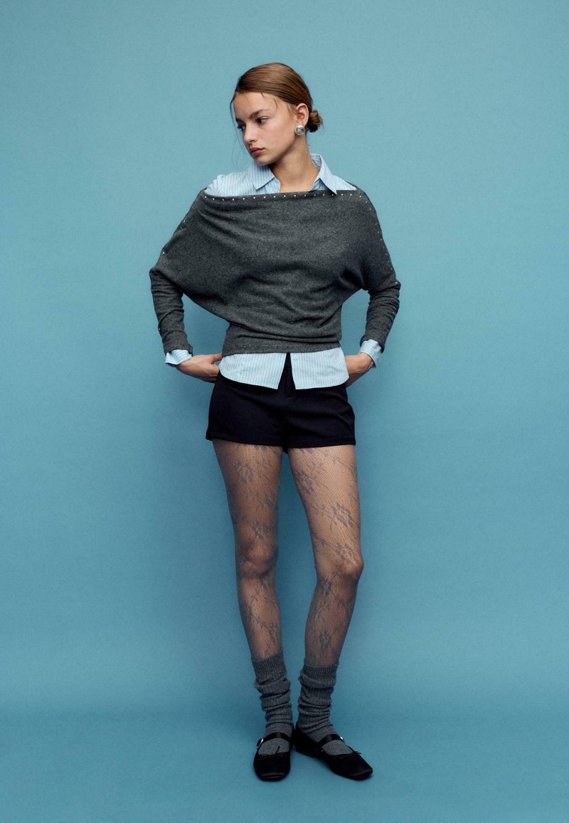 Siv prevelik pulover z modro črtasto srajco pod njim, črni kratki hlači, vzorčaste nogavice in sive nogavice do kolen s črnimi čevlji.