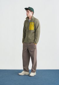 Νέος άνδρας με πράσινο καπέλο, σομόν φλις μπουφάν με κίτρινη τσέπη στο στήθος, καφέ παντελόνι και λευκά αθλητικά παπούτσια, στέκεται σε μπλε χαλί μπροστά σε λευκό τοίχο.