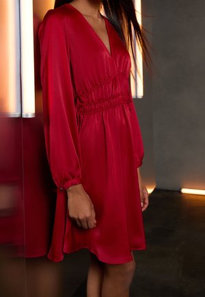 Femme vêtue d'une robe en satin rouge à manches longues, col en V et taille cintrée, debout près de lumières murales verticales dans une pièce peu éclairée.