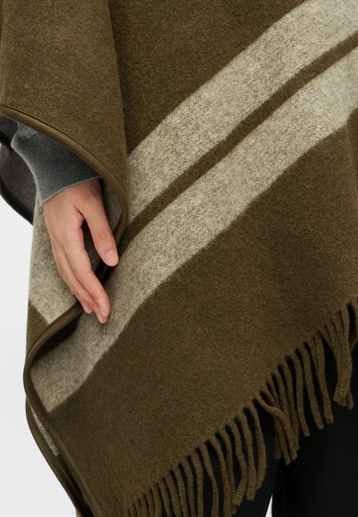 Mão descansando sobre um poncho de lã listrado em verde-azeitona e bege, com franjas, por cima de uma camisa de manga longa cinza e calças escuras.