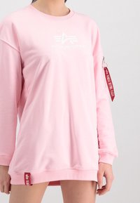 Jasnoróżowa, oversizeowa bluza wykonana z miękkiej tkaniny. Posiada okrągły dekolt, długie rękawy oraz zapinaną na zamek kieszeń z czerwoną metką "Remove Before Flight".