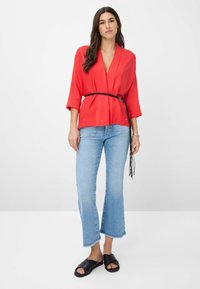 Rode, geribbelde wikkeltop met brede mouwen, om de taille cinched met een zwarte riem, gecombineerd met lichtblauwe cropped jeans en zwarte sandalen.