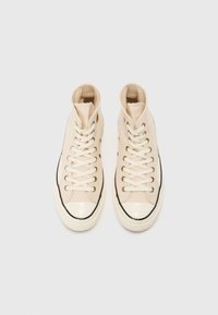 High-top sneakers in cream canvas, featuring white laces, gold eyelets, a black rubber sole, and a glossy toe cap.