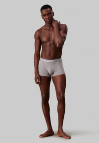 Sivi bombažni bokserki s bež logotipom Calvin Klein na pasu. Model stoji bos, prikazujejoč prilegajoč dizajn.