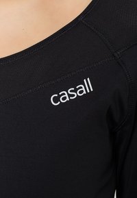 Top sportivo nero con scollatura rotonda, caratterizzato da cuciture piatte e un logo bianco "casall" sul lato. Tessuto liscio e elasticizzato.