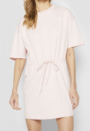 Žena v světle růžových krátkých šatech s tahem v pase, s malým bílým nápisem na horní části hrudi, s viditelnými tetováními na pažích.