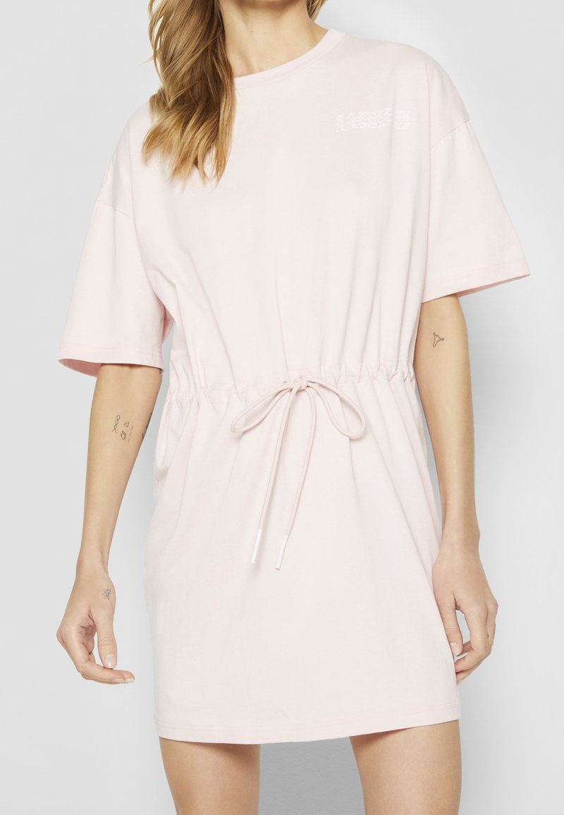 Femme portant une robe rose pâle à manches courtes avec une taille à cordon, comportant un petit texte blanc sur la partie supérieure de la poitrine, avec des tatouages visibles sur les bras.