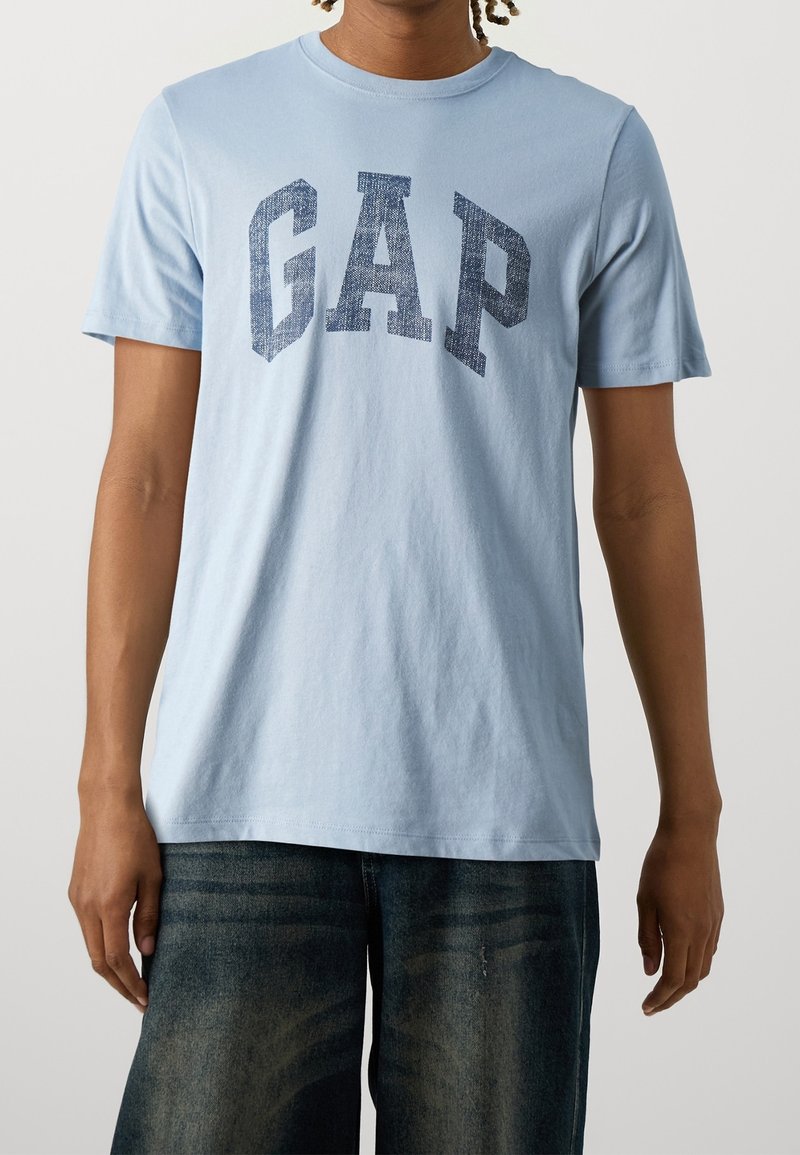 Personne portant un t-shirt à manches courtes bleu clair avec de grandes lettres bleu marine usées "GAP" sur la poitrine et un jean bleu foncé.