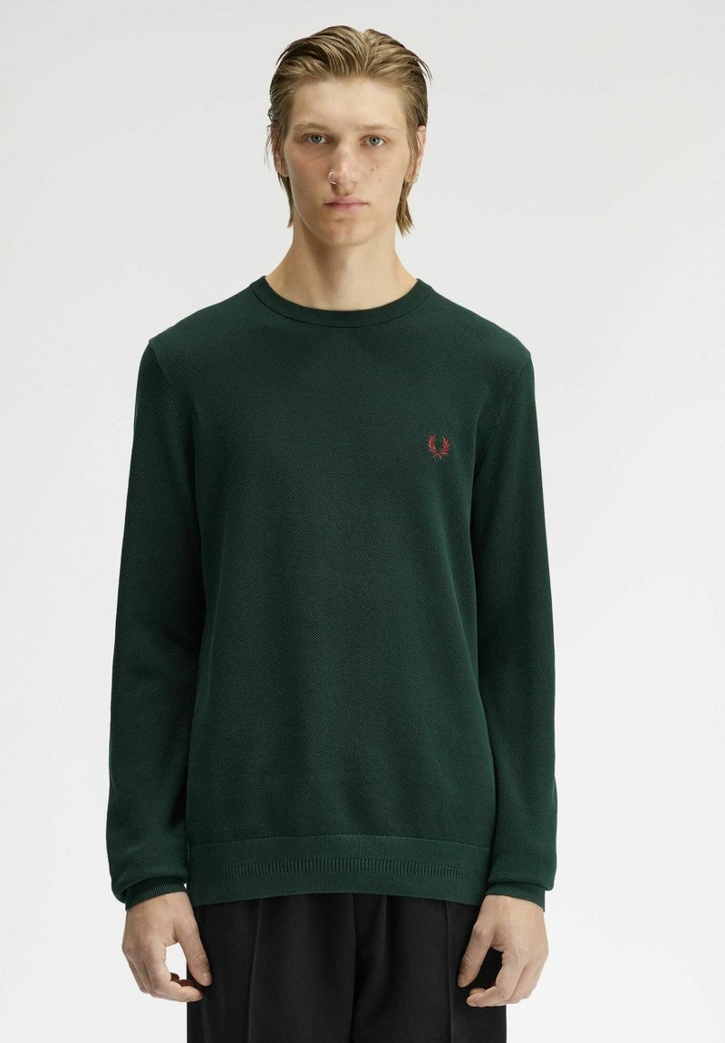 Modèle masculin aux cheveux clairs portant un pull à manches longues vert foncé et un pantalon noir, debout devant un fond clair uni.