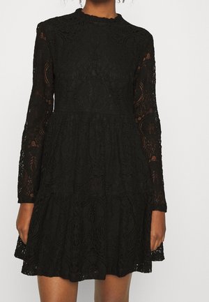 Robe en dentelle noire à manches longues, avec un design à volants et un col festonné. Motifs floraux détaillés et texture semi-transparente.