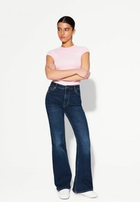 Hellrosa Kurzarmhemd, kombiniert mit blauen Schlagjeans, die eine hohe Taille und eine enge Passform an den Hüften aufweisen, abgerundet mit weißen Sneakern.