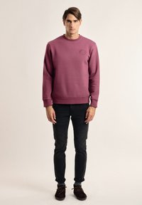 Sudadera burdeos con cuello redondo, puños y dobladillo de canalé; presenta un logo circular bordado en el pecho; combinada con jeans oscuros ajustados.