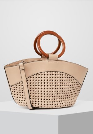 Beżowa torebka z plecioną dolną częścią, gładkim górnym wykończeniem, okrągłymi drewnianymi uchwytami oraz cienkim paskiem, spoczywająca na białym postumencie na tle szarej ściany.