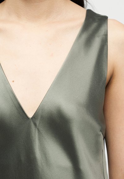 Prim-plan al unei persoane care poartă un top fără mâneci, din satin verde măsliniu, cu un decolteu adânc în V, care dezvăluie gâtul și partea superioară a pieptului.
