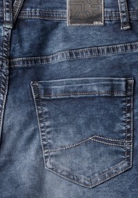 Tela de mezclilla en un color azul desgastado, que presenta un único bolsillo trasero con bordes cosidos y una etiqueta rectangular con un diseño en relieve.