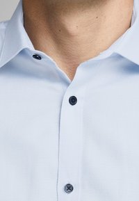 Närbild av en persons nacke och övre bröst som bär en ljusblå strukturerad skjorta med mörka knappar, kragen öppen upptill.