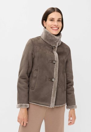 Eine lächelnde Frau, die eine braune Wildlederjacke mit großen Knöpfen, hohem Kragen und Schafsfellbesatz trägt, kombiniert mit hellbraunen Hosen.