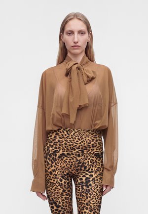 Femme aux cheveux blonds raides porte un chemisier marron transparent avec un grand nœud et un pantalon taille haute imprimé léopard, debout devant un fond uni.