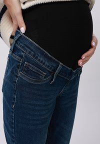 Trudnički traperice u tamnoplavom denimu, s rastezljivim pojasom za udobnost, s prednjim i stražnjim džepovima, prikazane na neutralnoj pozadini.
