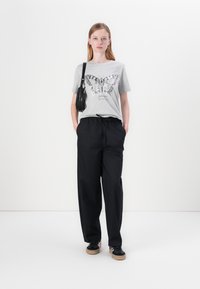 T-shirt grise avec un graphisme de papillon, pantalon noir ample et baskets noires avec semelle en caoutchouc. Le modèle porte un petit sac noir.