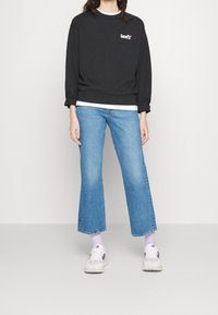 Sudadera negra con el logo blanco de Levi's, llevada sobre una camisa de manga larga blanca; combinada con jeans cortos de mezclilla de color azul claro y zapatillas blancas.