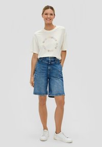 Crème t-shirt met meerkleurige cirkelvormige tekst, gecombineerd met blauwe denim shorts met een rechte snit en een rafelige zoom. Witte sneakers.