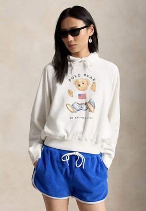 Vrouw met zwarte zonnebril, witte hoodie met teddyberen- en Amerikaanse vlagprint, blauwe shorts met trekkoord en schelpoorbellen.