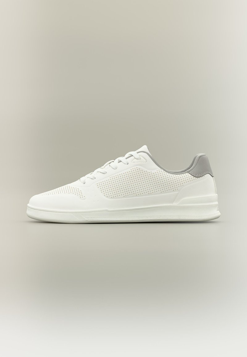 Baskets blanches basses avec détails perforés, languette grise au talon, semelle blanche et lacets blancs sur un fond neutre.