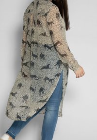 Blouse longue et transparente avec un imprimé animal texturé en noir et beige, présentant un ourlet asymétrique et des poignets boutonnés. Portée sur un jean bleu.