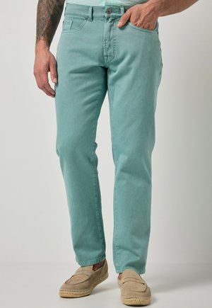 Mężczyzna noszący jasnozielone jeansy slim-fit i beżowe buty wsuwane, z jedną ręką w kieszeni, na neutralnym tle.