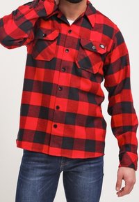 Camisa de franela a cuadros roja y negra con mangas largas, cierre de botones, dos bolsillos en el pecho y cuello clásico. Jeans de mezclilla visibles debajo.