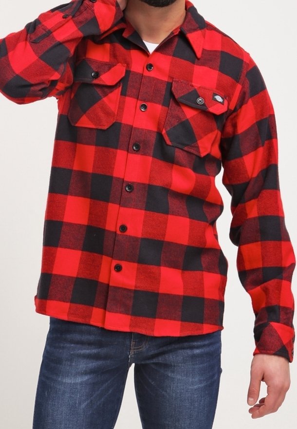 Camisa de franela a cuadros roja y negra con mangas largas, cierre de botones, dos bolsillos en el pecho y cuello clásico. Jeans de mezclilla visibles debajo.