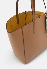 Tan læder tote taske med struktureret design, glat overflade og kontrasterende gul inderside. Har dual håndtag og et dekorativt mærke.