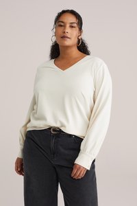 Pull à col en V de couleur crème avec un tissu texturé et des manches longues, associé à un jean taille haute foncé et une ceinture à accents dorés.