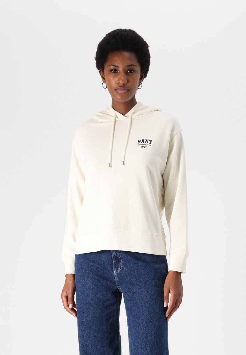 Kremowa bluza z kapturem, wykonana z miękkiego materiału, z przednią kieszenią typu kangur, sznurkiem do ściągania i logo "GANT 1949". Połączona z niebieskimi dżinsami.